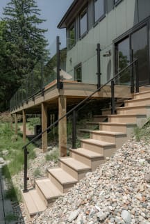 Black-post glass railing follows composite exterior stairs and an elevated deck on a hillside home.