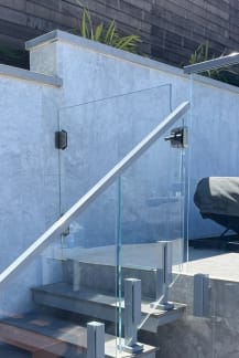 Clear glass panels on talon clamps with a stainless handrail follow an exterior stone stair by a pool deck.