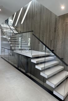 White floating treads with mid landing and frameless glass in a black base rail against a stone wall.
