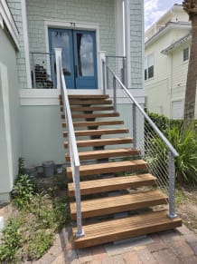 Wood floating treads on a mono stringer climb to a blue front door, framed by silver-post railing with horizontal infill.