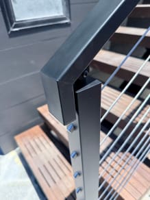 Close-up of a black square post and handrail elbow with stainless cable infill over wood stair treads.
