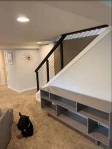 Black square-post cable railing with a dark wood handrail lines a basement staircase above a built-in shelf; a small dog sits nearby.