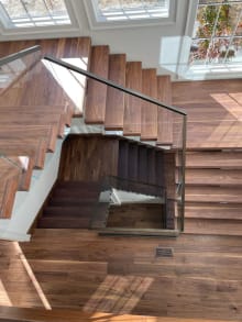 Overhead view of walnut stair treads and landing with glass railing and a black handrail, wrapping a multi-level stairwell.