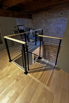 Black horizontal rod railing with a maple handrail surrounds a stair opening in a brick-walled loft.