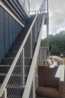 White posts and cable railing follow a long exterior stair along a blue-sided home above a patio.