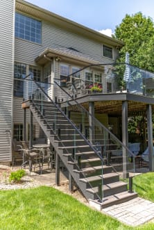 Tall exterior deck stair framed by frameless glass on black talon spigots leading to a covered patio and lawn.
