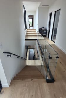 Base rail glass and a black wall handrail lead down an oak stair from a long hallway