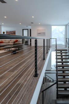 Black cable railing with a round top rail lines a balcony above a floating stair and living room.