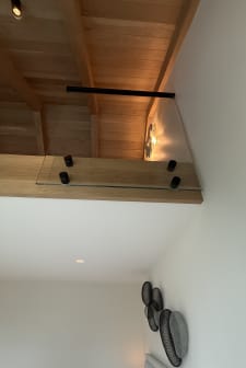 Black standoff pins fix a glass guard to the oak fascia of a timber-ceilinged loft above a bedroom.