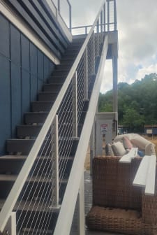 White posts and cable railing follow a long exterior stair along a blue-sided home above a patio.