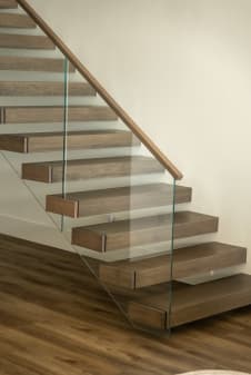 Thick walnut-tone cantilever treads float off a white wall beside a frameless glass guard with a wood handrail.