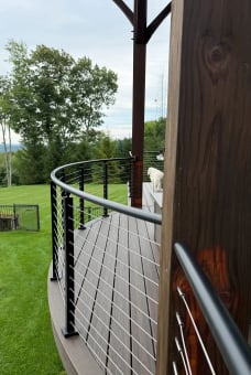 Black curved cable railing follows the edge of a round composite deck beside a timber post.