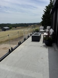 Frameless glass panels on black spigots line a concrete patio with a fire table overlooking pastures.