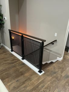 Black perforated metal panels with a dark wood cap guard a stair opening on a wood floor.