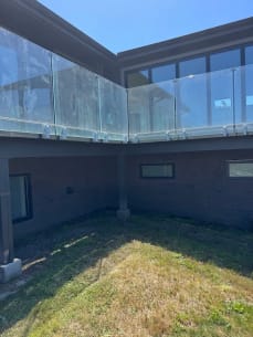 Frameless glass on side-mount brackets wraps an elevated deck on a gray modern home above a lawn