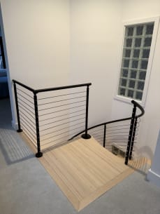 Black cable railing guards the upper opening of a maple spiral stair beside a glass block window.