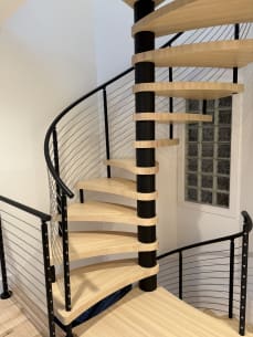 A maple spiral stair winds around a black column, wrapped by a curved black cable railing.