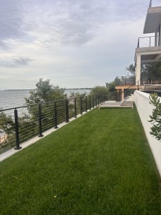 Black cable railing runs along a cliff-top lawn above the water beside a modern home.