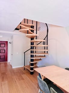 Black-column spiral staircase with light wood treads and a curved black cable railing beside a dining table.