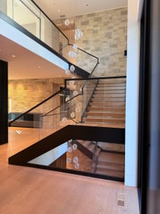 Black-framed floating stair with light oak treads and frameless glass railing wraps around a stone wall with a hanging glass-orb chandelier.