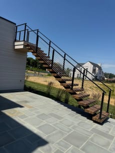 Hardwood floating treads on a black mono stringer climb a white home's exterior to an upper deck, edged by black cable railing.