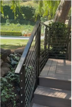 Bronze-black posts with horizontal cable infill follow composite deck steps down to a shaded garden.