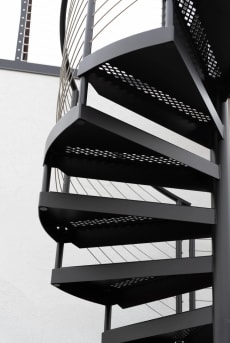 Perforated black steel spiral treads climb beside vertical cable infill against a white wall.