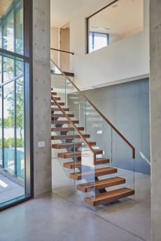 Floating wood treads on a center stringer with frameless glass and a wood handrail beside a concrete column and tall windows.