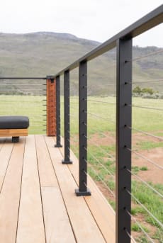 Black side-mounted posts with cable infill and black top rail along a light wood deck facing open hills.