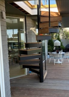 Black steel exterior spiral stair with IPE wood treads descending to a wood deck beside a shingled home.