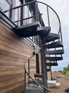 Gray steel spiral stair with curved cable railing rises along a wood-clad home to an upper deck near the ocean.