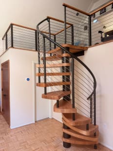A wood-tread spiral stair with a curved black rail and horizontal infill rising to a cable-railed loft.