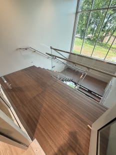 A wood landing with glass railing and stainless handrails beside a tall window in a commercial building.