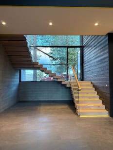 Light wood stacked treads with LED-lit nosings rise along a dark wood wall beside glass railing and a tall window.