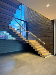 Boxy wood stacked stair with glass railing and wood handrail climbs a dark timber wall to a window-lit landing.