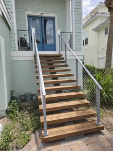 Wood floating treads on a mono stringer climb to a blue front door, framed by silver-post railing with horizontal infill.