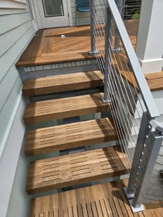 Looking down slatted hardwood floating treads beside a silver cable railing post to an IPE deck landing.