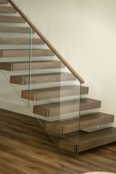 Thick walnut-tone cantilever treads float off a white wall beside a frameless glass guard with a wood handrail.