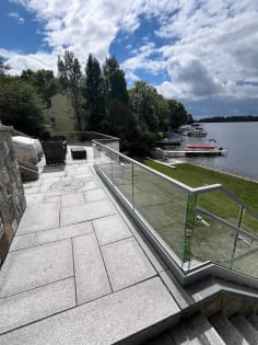 Frameless glass railing on a gray aluminum base rail edges a granite patio overlooking a lake and boat docks