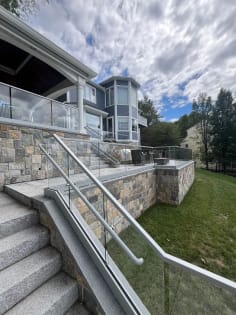 Glass base rail railing with a round handrail climbs granite steps beside a stone wall under a cloudy sky