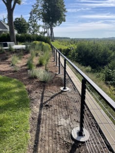 Black surface-mount cable railing runs along a landscaped hillside walkway with a wooded valley view