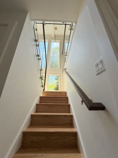 A walnut wall-mounted handrail lines an enclosed oak stair rising toward glass railing and tall windows.