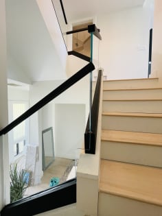 Frameless glass railing in a black channel with a square black handrail follows a light oak switchback stair.