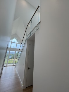 Frameless glass panels on white standoff pins with a wood handrail rise along an oak stair toward a gabled window wall.