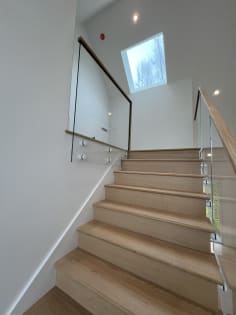 An oak staircase climbs toward a skylight between glass railings on standoff pins with wood handrails.