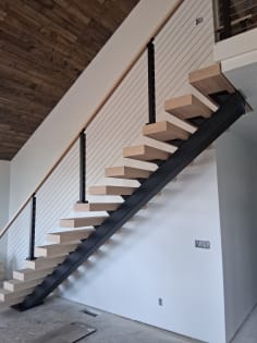 A FLIGHT Mono stair with white oak treads and black-post cable railing rises to a loft under a rustic wood ceiling.