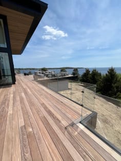 Frameless glass railing wraps the edge of a long hardwood deck overlooking evergreens and water.
