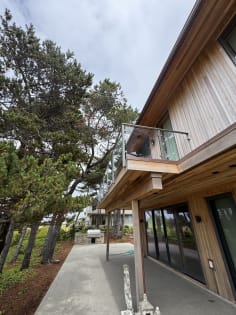 Glass panels between slim gray posts with a top cap line a second-story balcony on a cedar-clad home among pines.