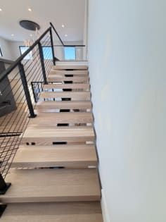 White oak floating treads rise along a white wall beside a black cable railing.
