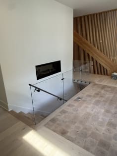 Frameless glass railing with black handrail around a stair opening near a linear fireplace and slat wall.